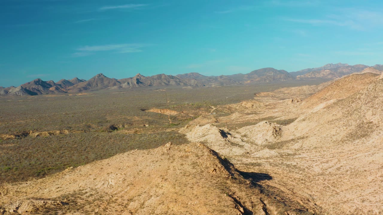 Wide aerial view near Cabo Pulmo, Baja California Sur, showing a sandy foreground ridge, scrub desert plain, distant mountains, blue sky, and no visible people.