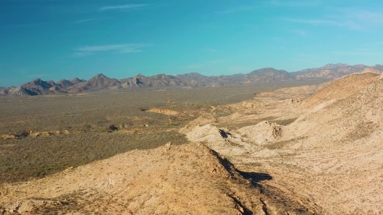 Aerial desert frame near Cabo Pulmo with eroded sandy washes in the foreground, muted green scrub across the plain, distant mountain ranges, and no visible people.