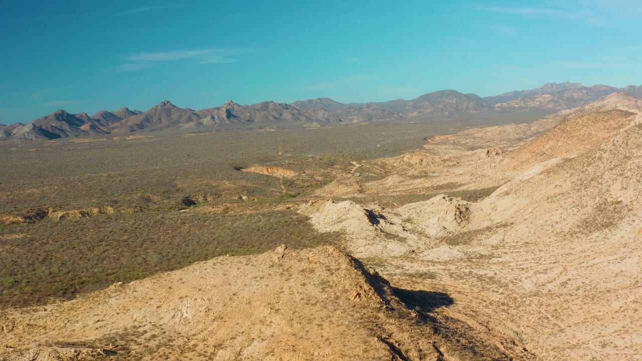 Cabo Pulmo desert view with pale foreground ridges, a broad scrub plain, distant mountain range, clear blue sky, and no visible people at the opening frame.