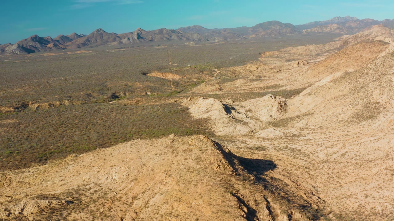 Aerial view one second into the Cabo Pulmo clip, showing the foreground ridge dropping toward pale dry washes, scrubby desert flats, distant mountains, and no visible people.