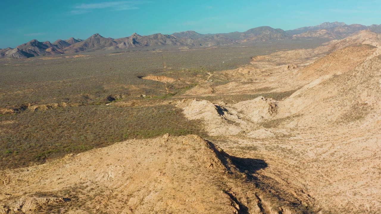 Midwide aerial frame over Cabo Pulmo with pale rocky slopes at right, darker scrubland across the basin, a mountain line in the distance, and no visible people.