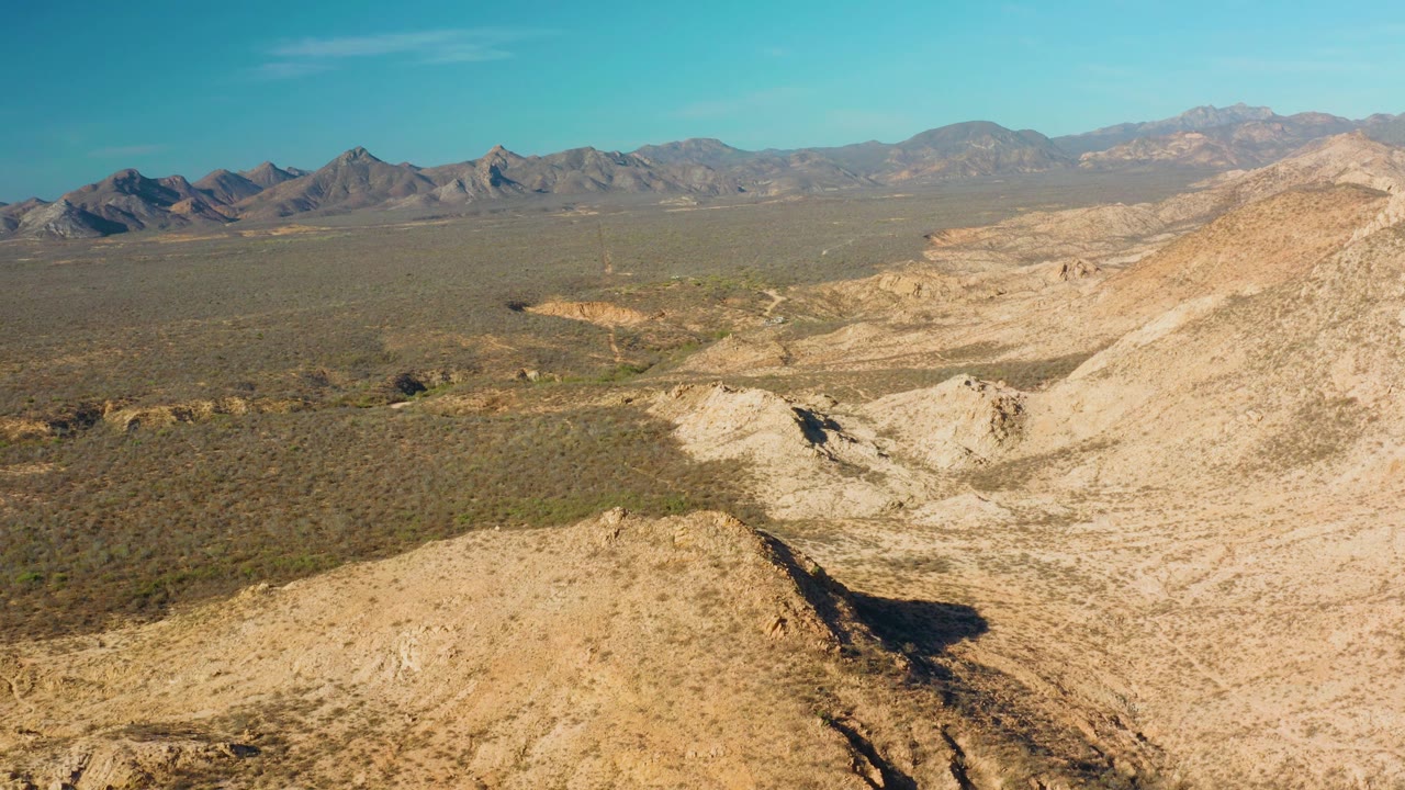 Aerial frame from Cabo Pulmo showing the foreground hill crest, a shadowed cut in the dry terrain, broad scrub flats, distant mountain ridges, and no visible people.