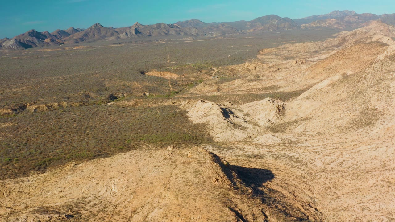 Aerial view in Cabo Pulmo showing a sandy foreground hill with a dark shadow edge, pale dry wash, scrub plain, mountain horizon, and no visible people.