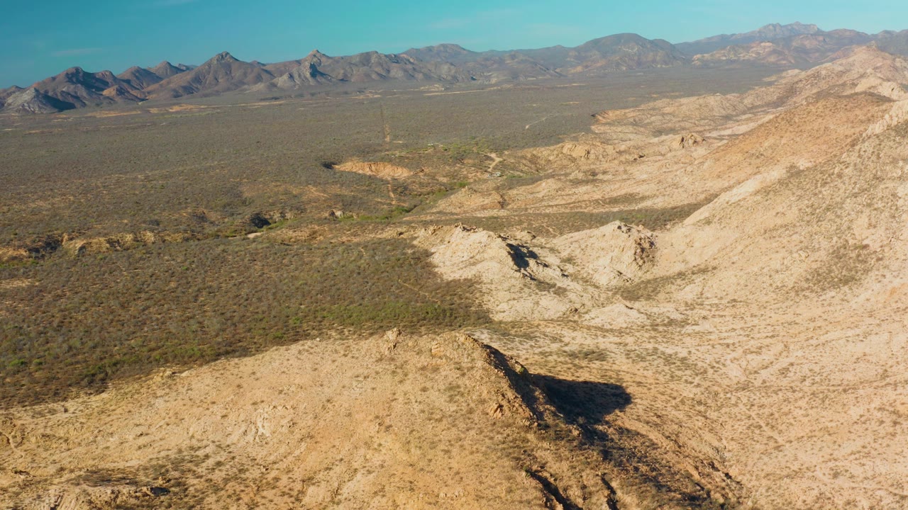 Cabo Pulmo aerial frame with tan ridges in front, a pale drainage channel through the center, darker desert scrub beyond, mountains on the horizon, and no visible people.