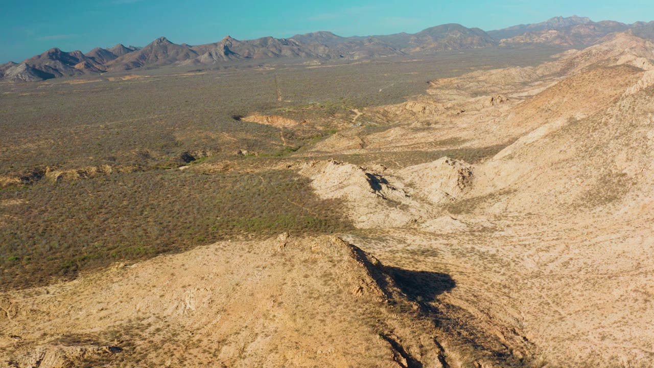 Mid-clip aerial view over Cabo Pulmo arid terrain, with a pale hillside crossing the foreground, scrub-filled flats, distant mountain ridges, blue sky, and no visible people.