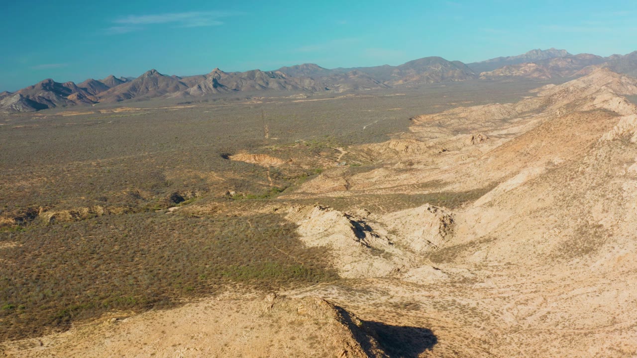 Aerial frame of Cabo Pulmo desert with pale rocky hills at right, broad brushy valley at center, mountains across the horizon, and no visible people.