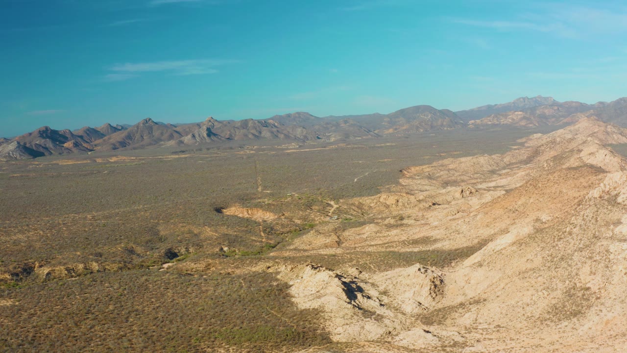 Sunlit desert valley with pale ridge in the foreground and mountains beyond.