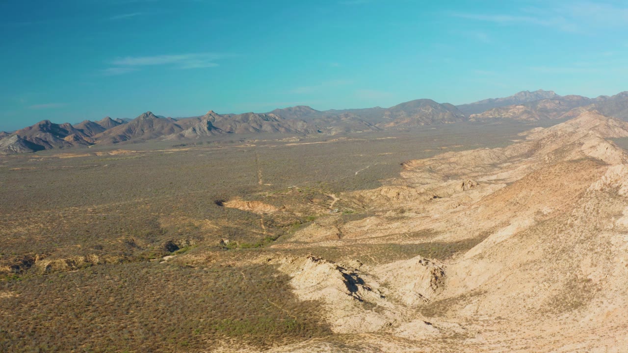 Cabo Pulmo aerial frame with rocky slope and eroded light-colored terrain in the foreground, open desert valley beyond, mountain ridges across the horizon, and no visible people.
