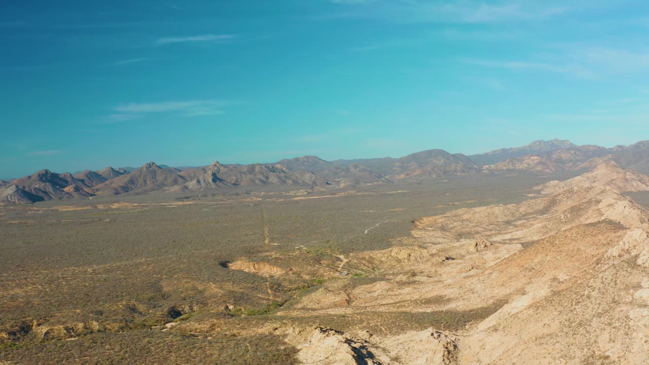 Opening frame of Cabo Pulmo desert terrain with a pale rocky ridge in the foreground, flat scrub plain beyond, distant mountains on the horizon, and no visible people.