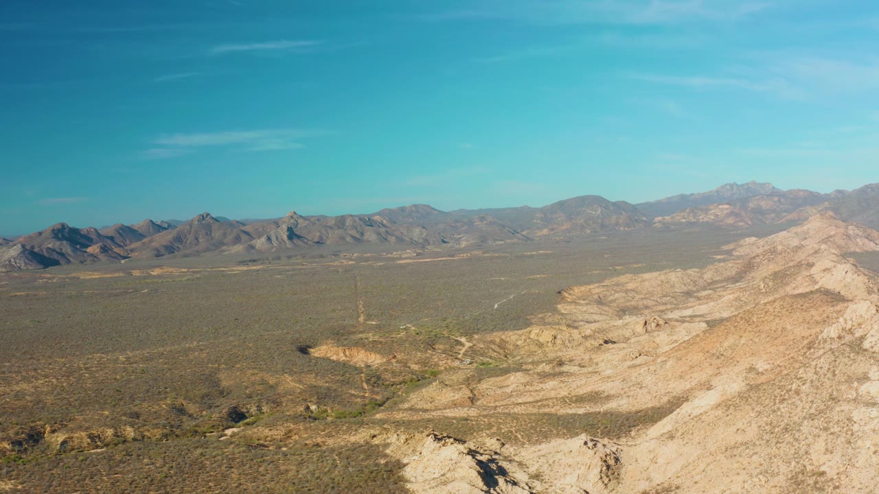 A wide Cabo Pulmo landscape shows tan rock in the foreground, darker scrubland across the valley floor, distant ridges, and no visible people under a blue sky.