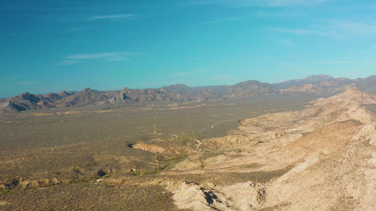 Mid-clip view over Cabo Pulmo desert shows a broad plain, rough foreground rock, a faint track or wash through the terrain, distant mountains, and no visible people.