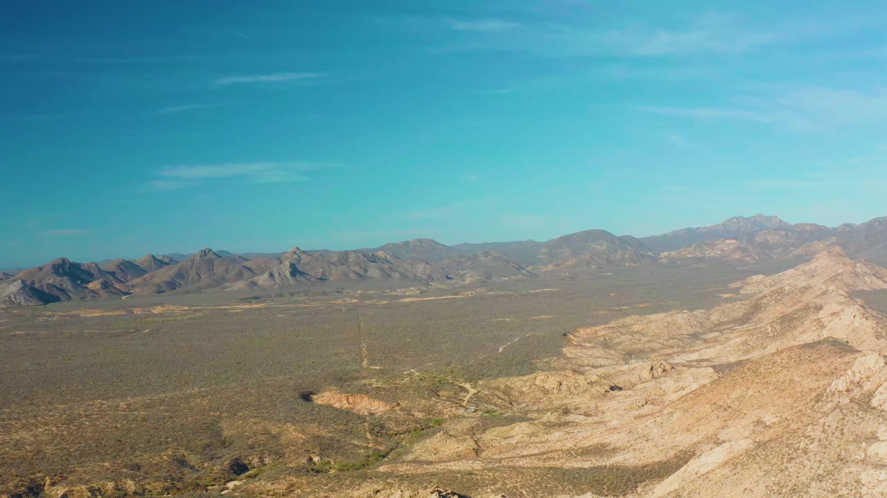 Aerial frame over Cabo Pulmo showing a light tan rocky slope at right, low desert vegetation across the middle ground, layered mountains in the distance, and no visible people.