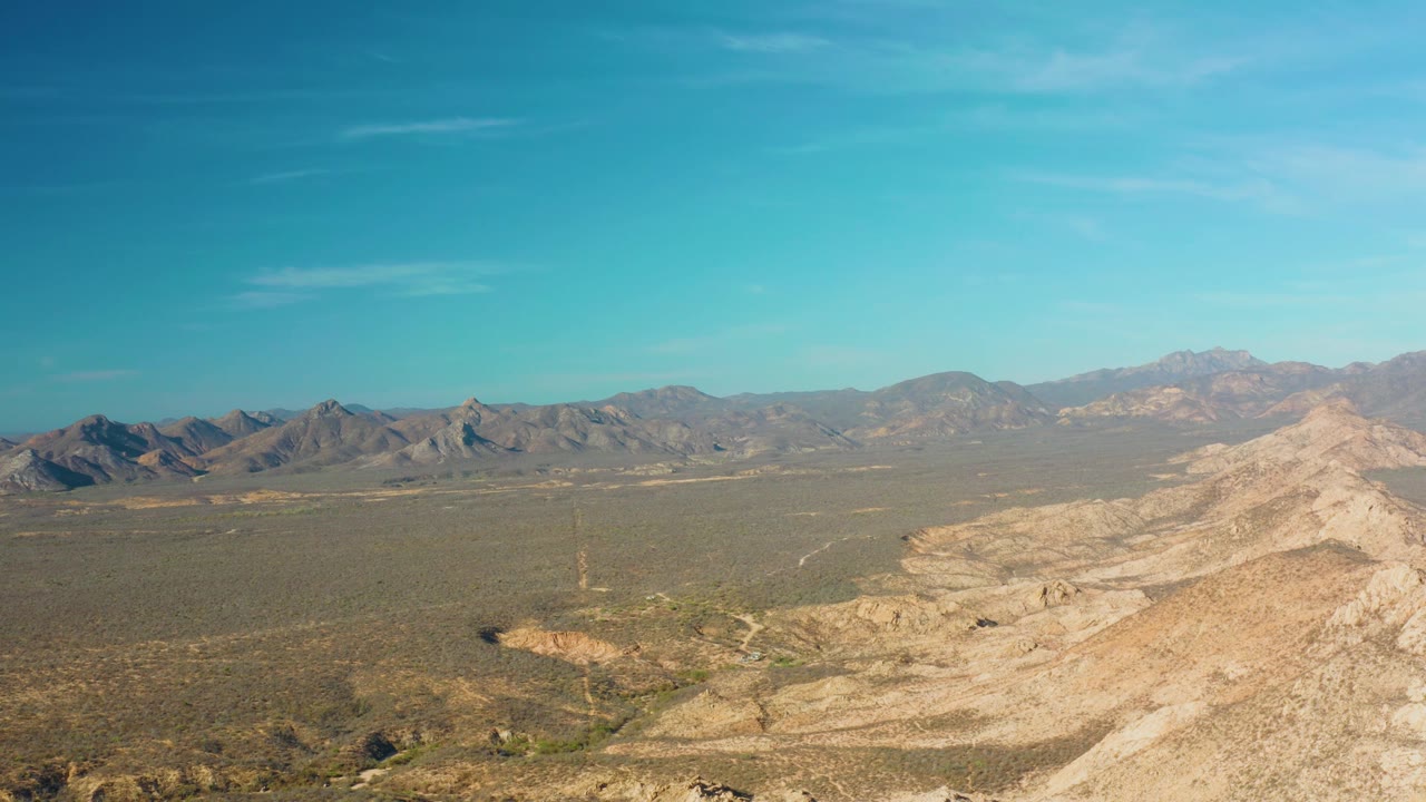 Aerial view in Cabo Pulmo with rough foreground rocks, scattered desert shrubs, a broad flat basin, rugged hills and mountains beyond, and no visible people.