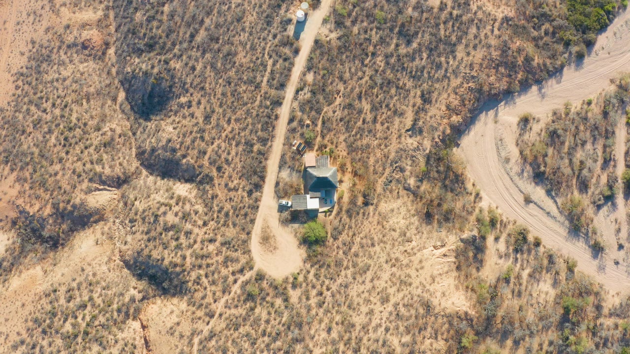 Overhead view in Cabo Pulmo, Baja California Sur, showing a pale dirt road beside dry scrub, a small roofed structure, a nearby tank, a vehicle on the road, and no visible people.