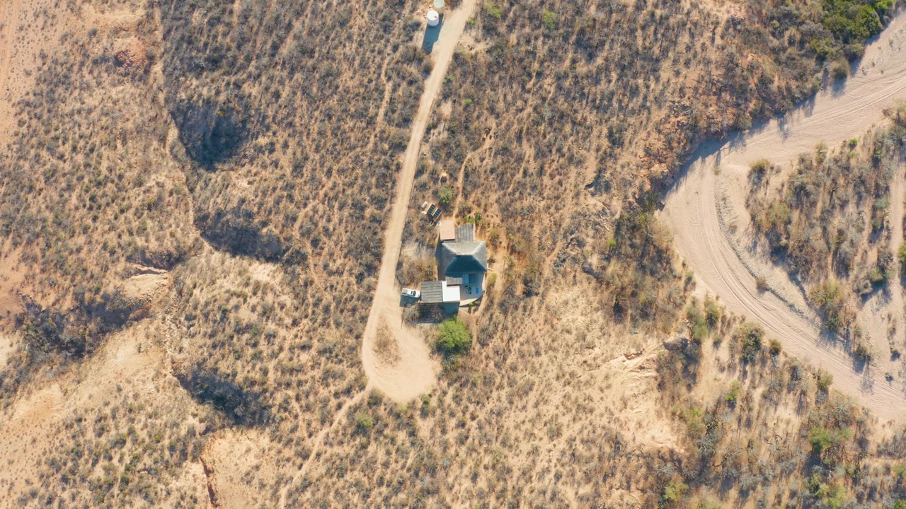 Top-down view in Cabo Pulmo, Baja California Sur, showing the vehicle continuing along the narrow dirt road past a house-like structure, dry grass patches, and no visible people.