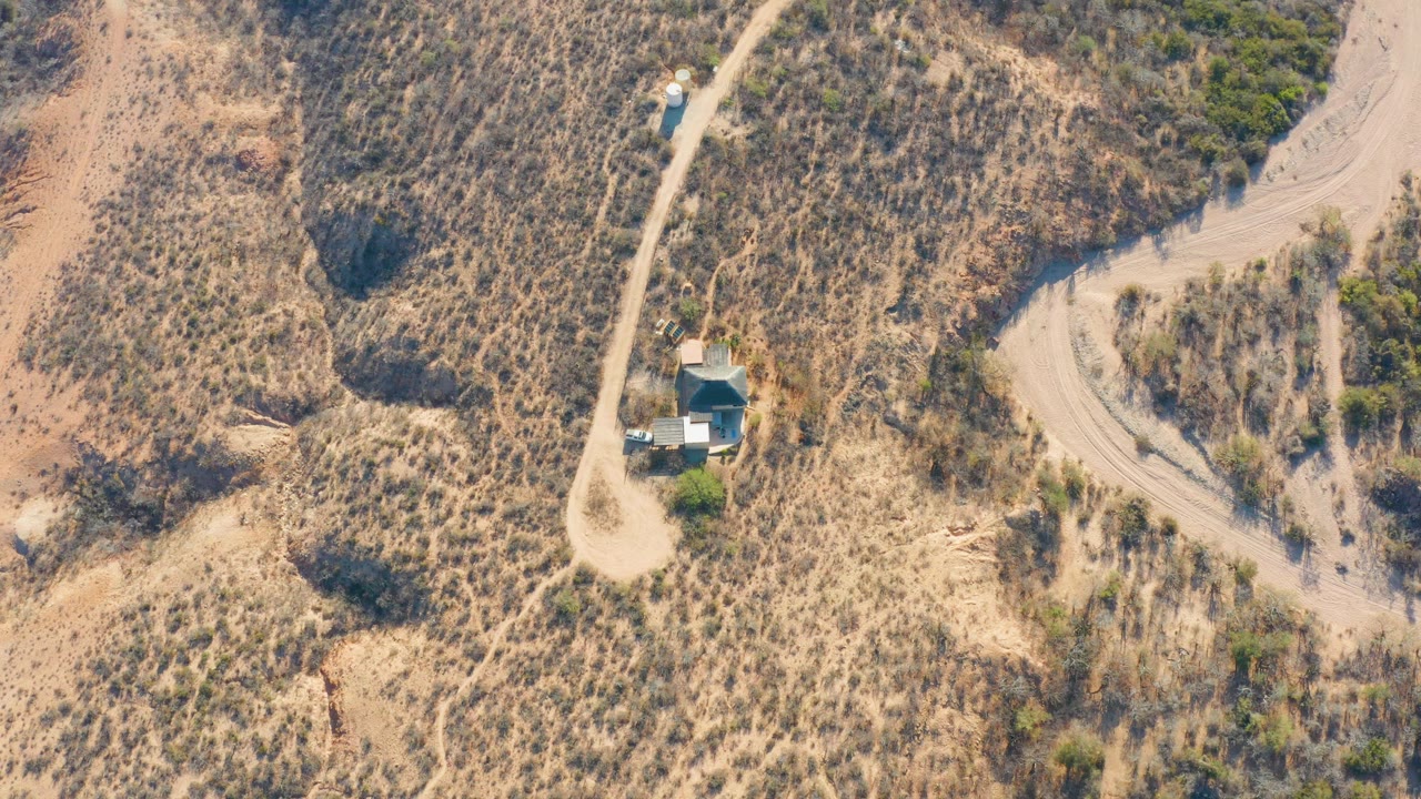 High aerial frame in Cabo Pulmo, Baja California Sur, Mexico, showing a pale dirt road through dry scrub, a small blue-roofed structure, a blue vehicle nearby, and no visible people.
