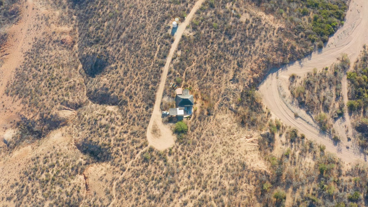 Aerial view of a blue vehicle beside a small structure along a winding dirt road in dry terrain.