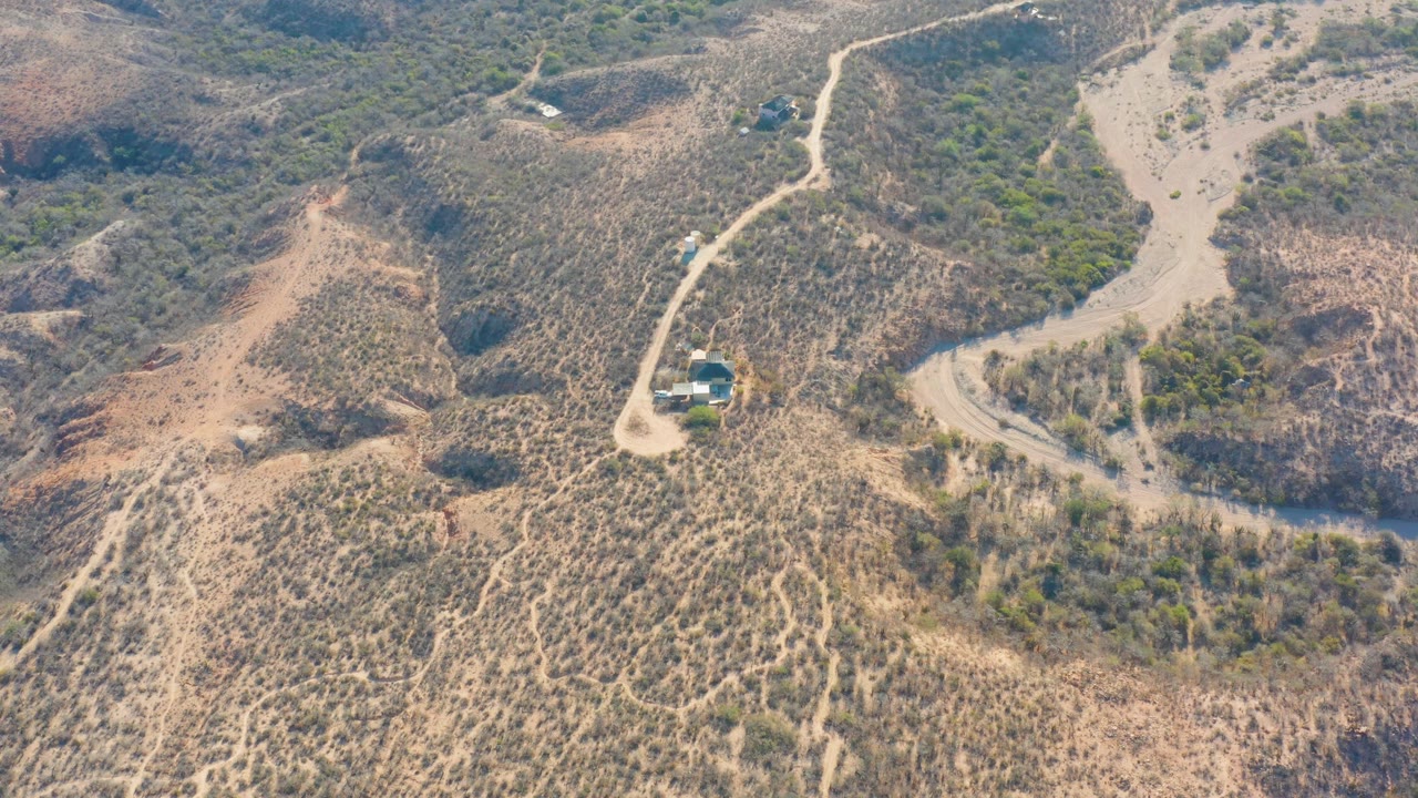 Aerial view in Cabo Pulmo with a pale winding road crossing dry terrain, scrub patches on the hills, a small building near the road, and no visible people.