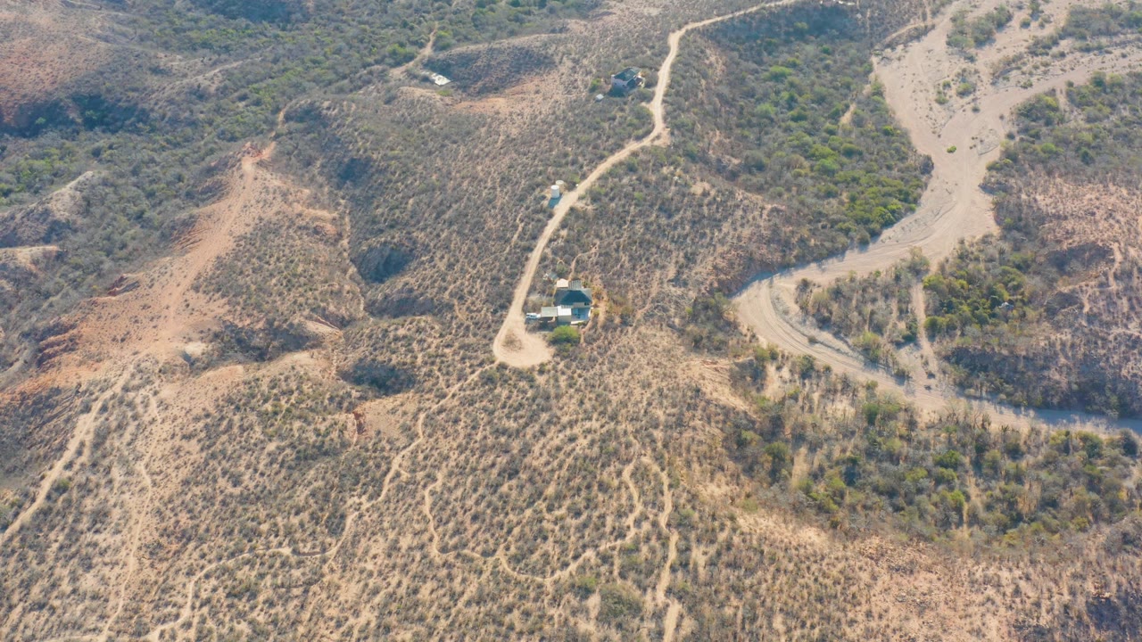 High aerial frame over Cabo Pulmo showing a vehicle on the curving dirt road, dry ridges, scattered green shrubs, and no visible people.
