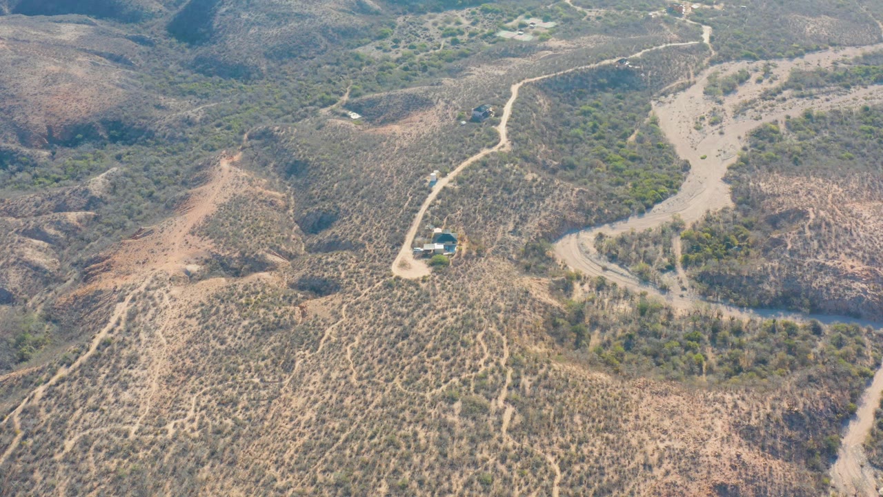 High aerial view in Cabo Pulmo, Baja California Sur, showing a small vehicle near a bend in a pale dirt road through dry scrub hills, with no visible people.