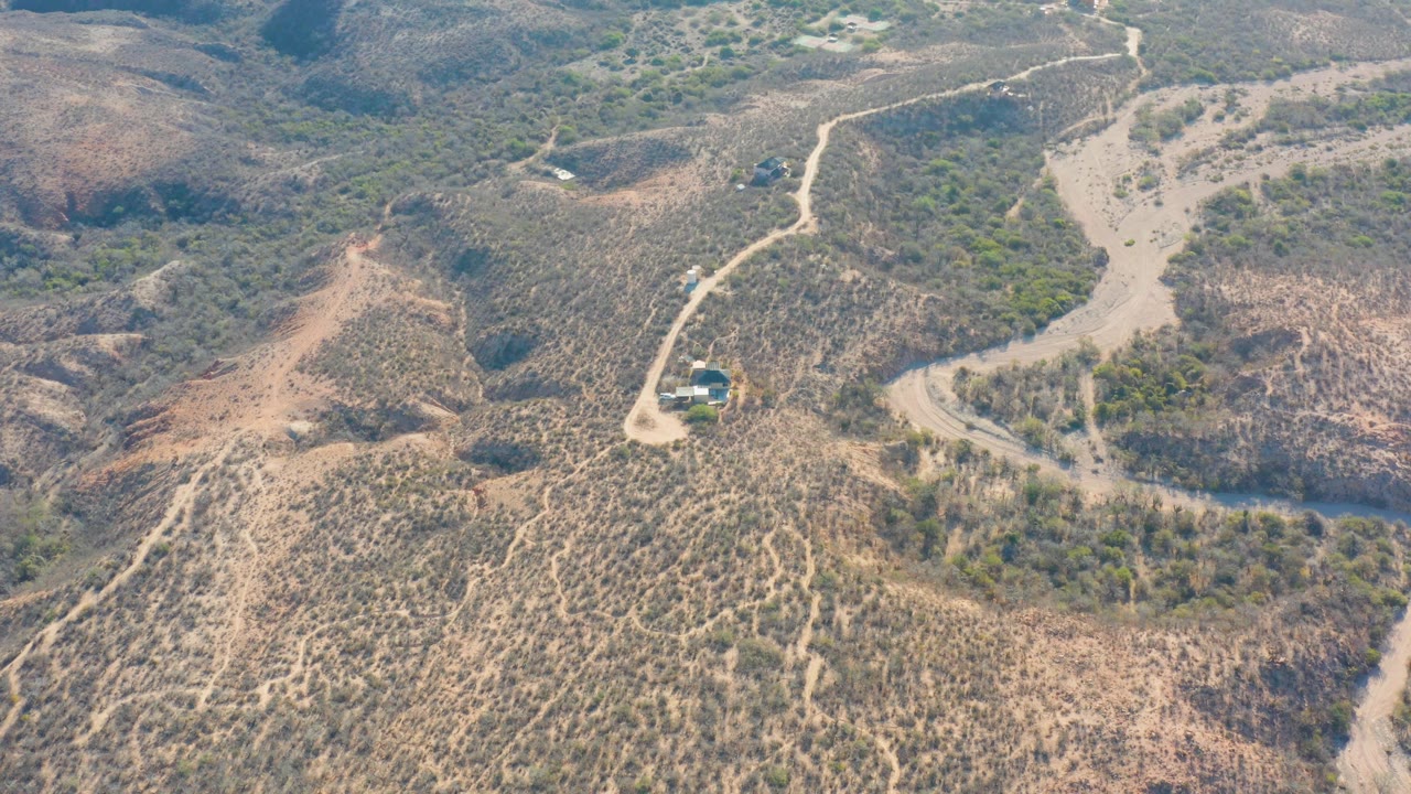 High drone view over Cabo Pulmo terrain, with the car close to a curved road section and dry ridges rising around sparse green vegetation, with no visible people.