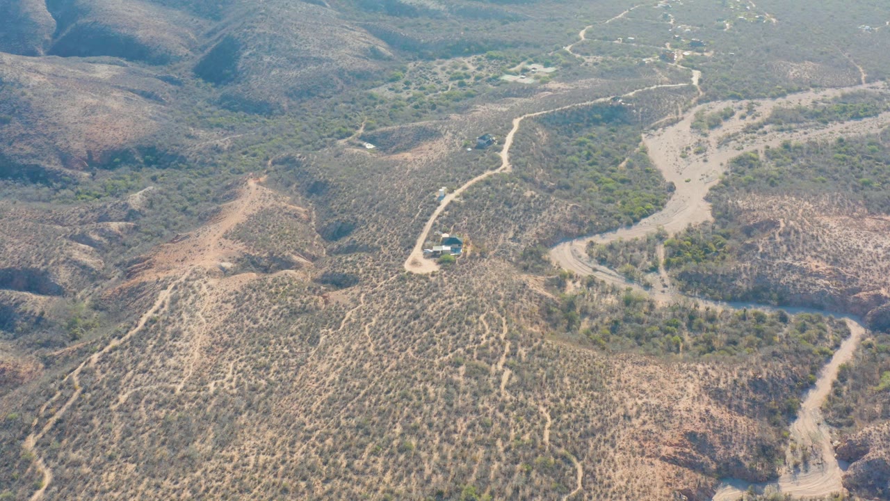 Elevated Cabo Pulmo view with a vehicle on the lower road, scrubby green patches around a dry wash, and blue-gray hills in the distance, with no visible people.