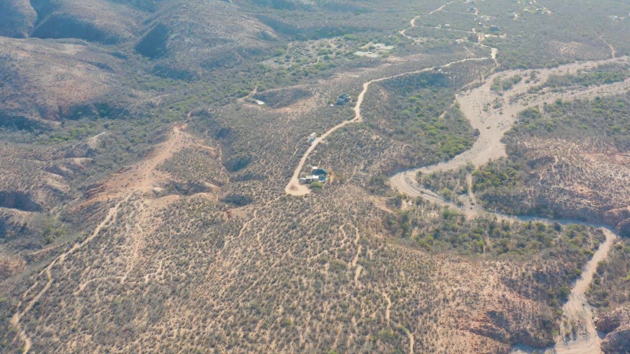 Drone-like aerial frame over Cabo Pulmo terrain, where a small vehicle sits on the lower-left dirt road below a broad dry hillside and scattered vegetation, with no visible people.