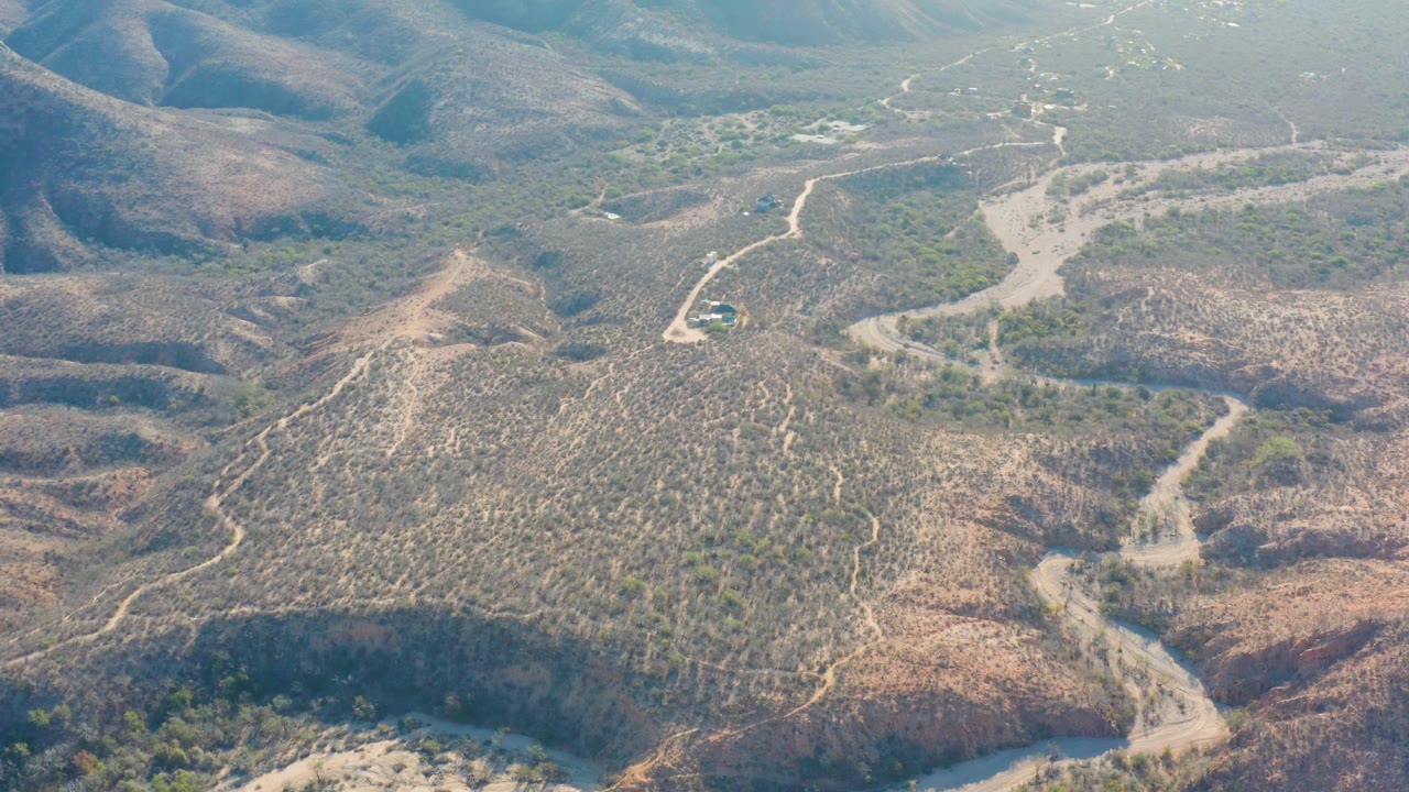 Aerial frame in Cabo Pulmo showing a small vehicle beside a curved dirt road, dry brush-covered hills, pale washes, and no visible people.