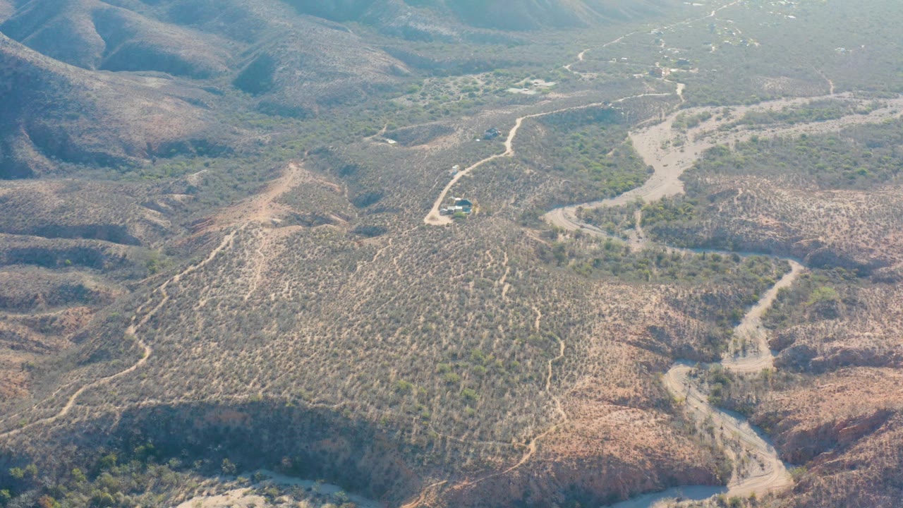 Drone-like aerial view of Cabo Pulmo terrain with the road shifting across the center, scrubby tan hills, distant ridgelines, and no visible people.