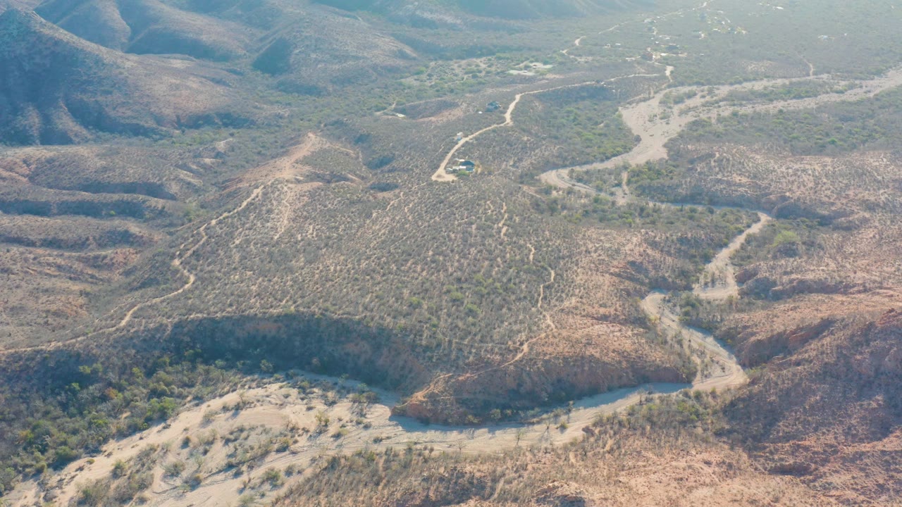 High aerial frame in Cabo Pulmo, Baja California Sur, showing a small vehicle on a pale dirt road amid scrub-covered hills and no visible people.