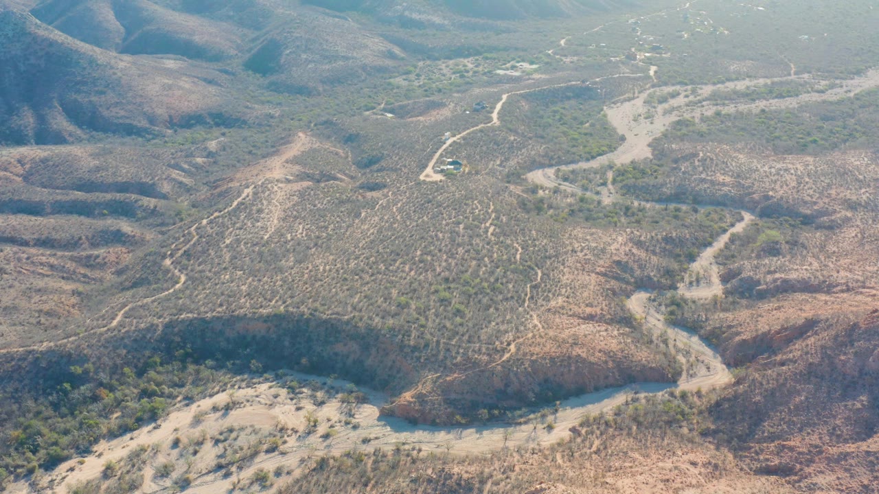 Aerial frame over Cabo Pulmo, Baja California Sur, with the dirt road bending through brown rocky hills, patches of green vegetation, and no visible people.