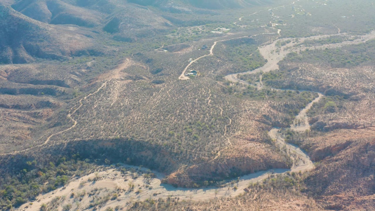Cabo Pulmo aerial view with a dirt track descending between dry slopes, scrub vegetation, a tiny vehicle near the road, and no visible people.