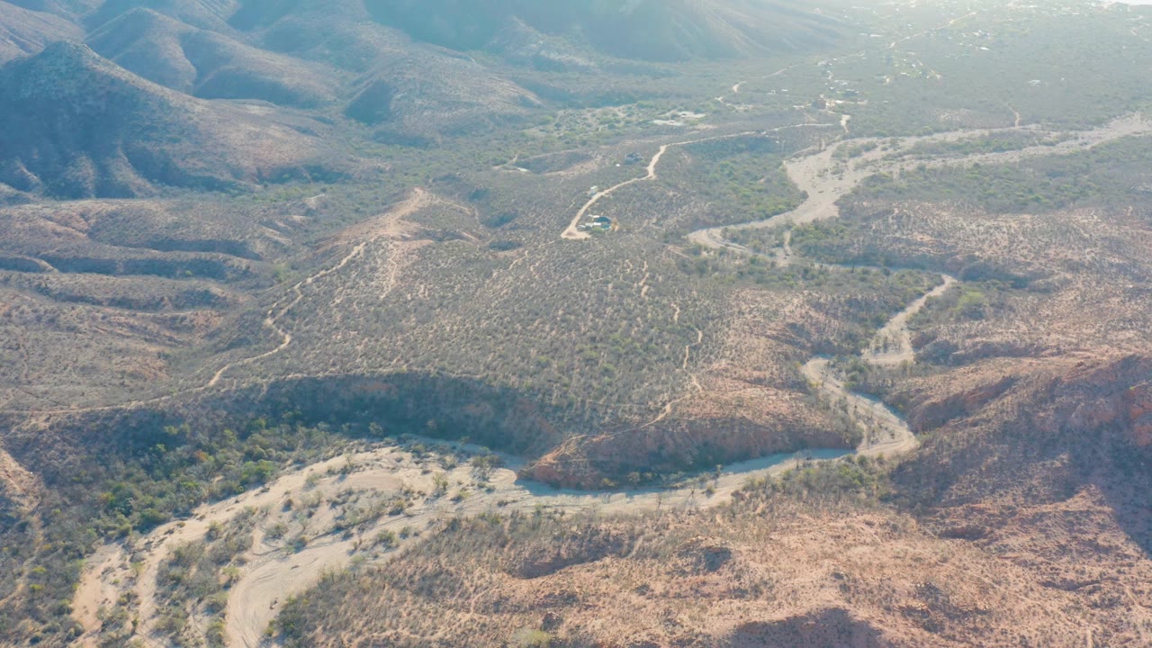 High aerial view in Cabo Pulmo with a dusty road bending through scrubby brown slopes, pale wash channels, hazy ridges, and no visible people.
