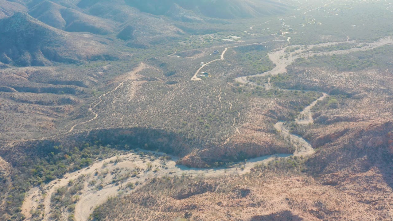 Aerial view over Cabo Pulmo showing small vehicles on a pale dirt road, open arid valley ground, darker green vegetation pockets, distant mountains, and no visible people.