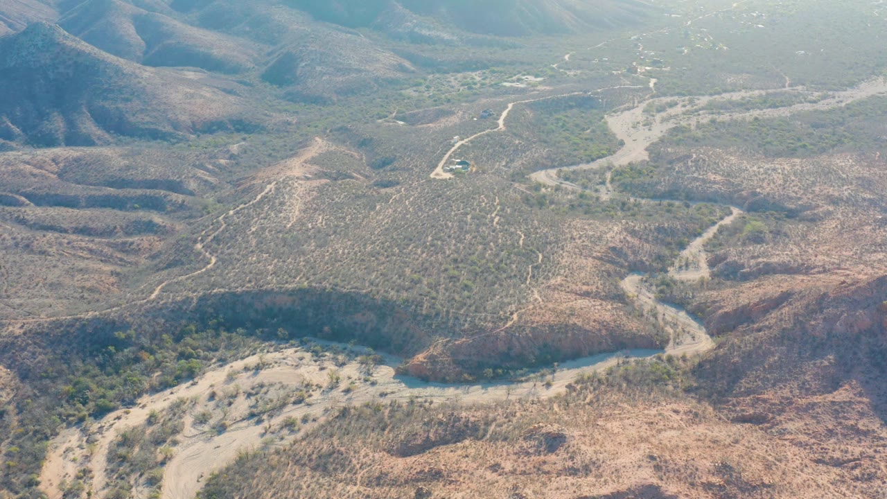 A Cabo Pulmo aerial frame with a winding dirt road, branching wash or riverbed, dry brush, shadowed hill contours, and no visible people.
