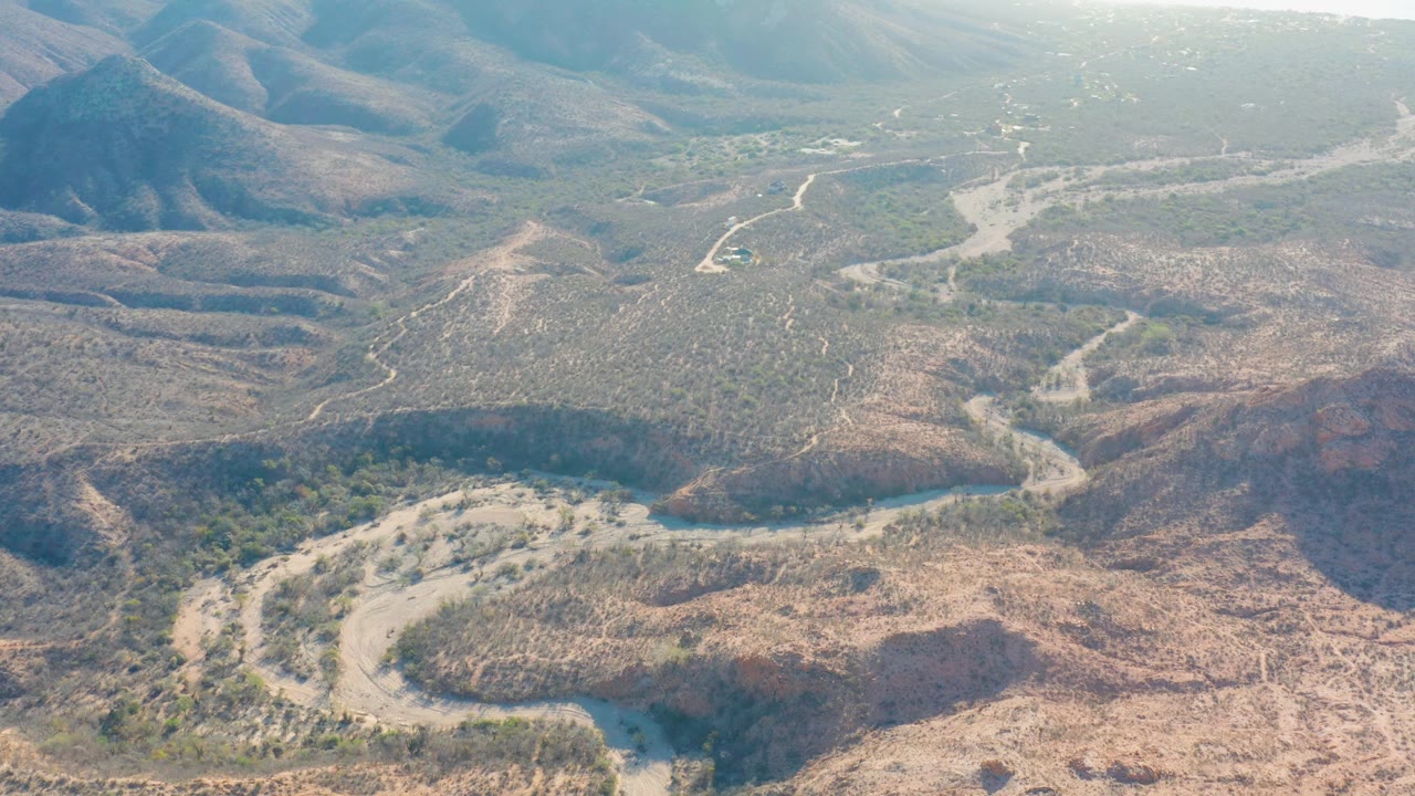 Aerial view in Cabo Pulmo, Baja California Sur, with the river channel crossing the lower frame and scrub-covered hills stretching toward a bright hazy horizon, no visible people.