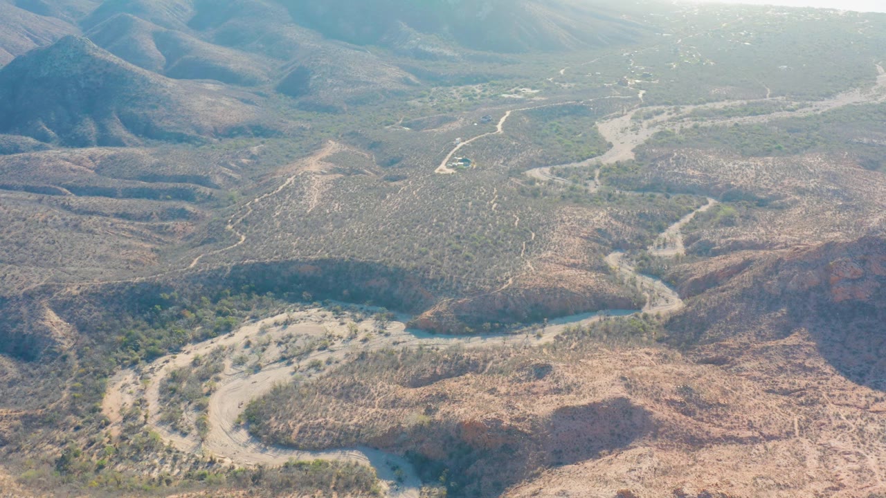Cabo Pulmo aerial frame showing a meandering wash between reddish-brown ridges, scattered green shrubs, and pale tracks across the valley, with no visible people.