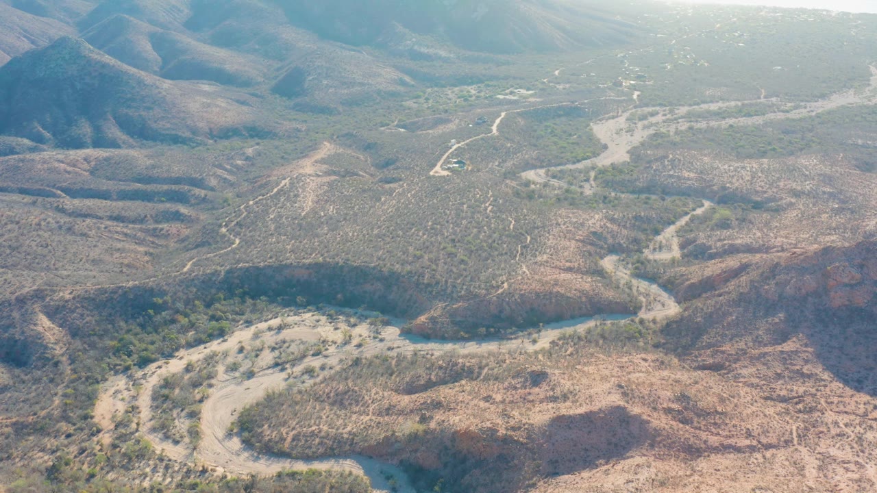 Aerial frame over Baja California Sur with a curved pale riverbed in the foreground, darker green vegetation near the bend, and hazy mountain slopes beyond, no visible people.