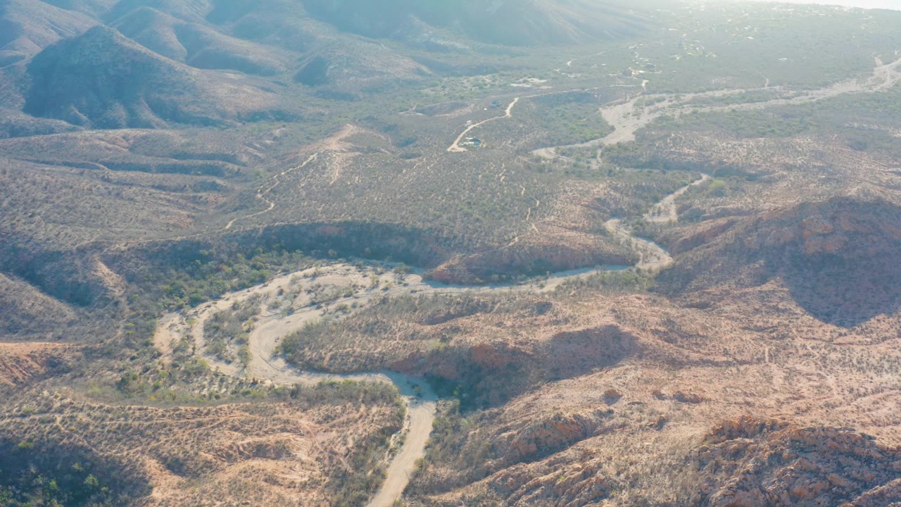 Aerial frame from Cabo Pulmo showing tan hills, scattered green scrub, a pale road or wash bending across the foreground, distant mountains, and no visible people.