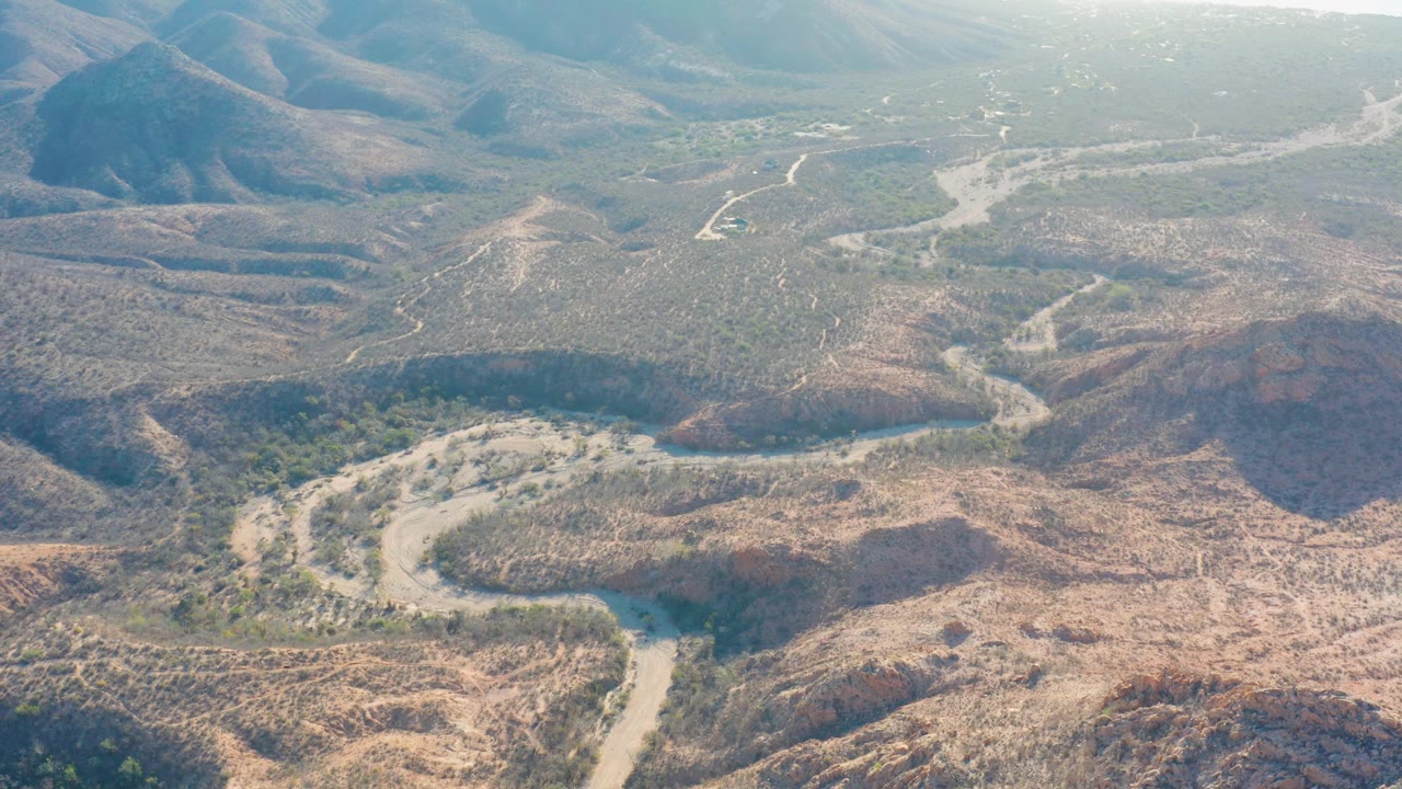 Wide drone view over Cabo Pulmo dry terrain with a winding dirt route in the lower frame, layered low hills, muted green vegetation, and no visible people.