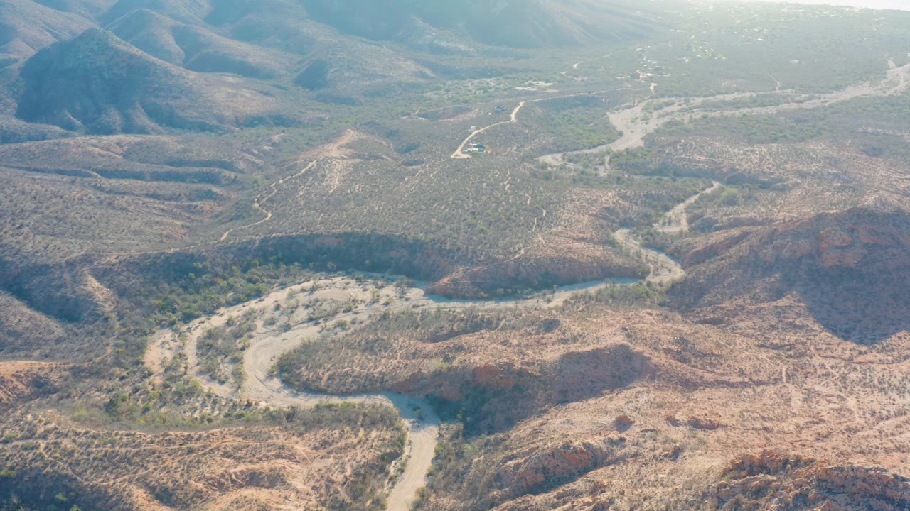 High view across Cabo Pulmo desert valley terrain, with pale tracks threading through sparse scrub, hills rising at midframe, distant mountains, and no visible people.