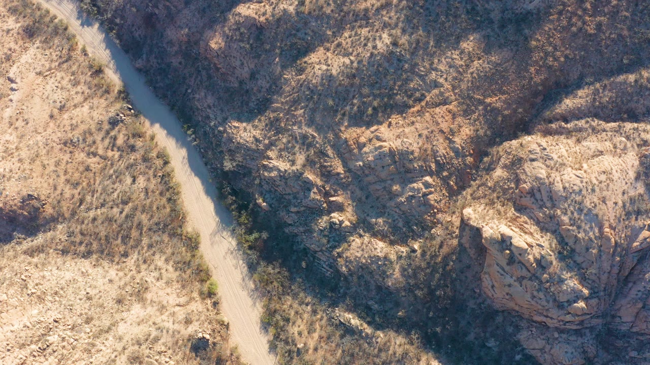 Aerial frame from Cabo Pulmo, Baja California Sur showing a pale dirt road along the left edge, dry scrub, rocky beige slopes, dark ravine shadows, and no visible people.