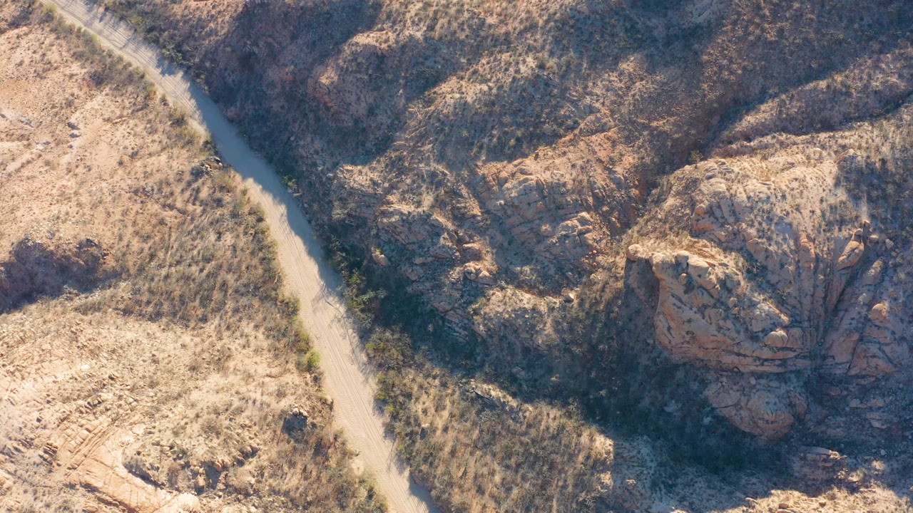 Overhead view in Cabo Pulmo, Baja California Sur, showing a narrow pale dirt road along dry scrub and dark rocky ground, with no visible people.
