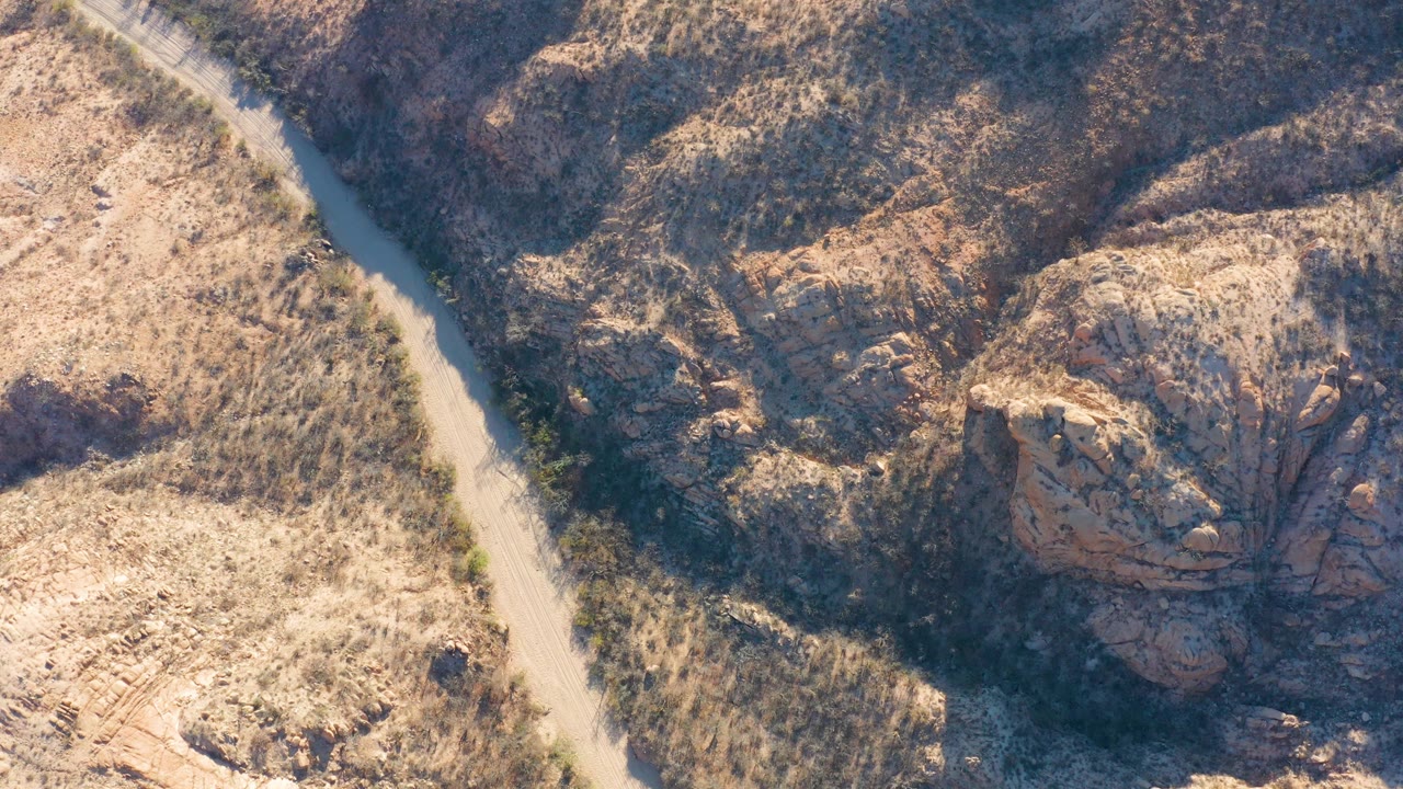 High overhead view in Cabo Pulmo, Baja California Sur, with a pale dirt track beside dry brush and shadowed rock cuts, with no visible people.