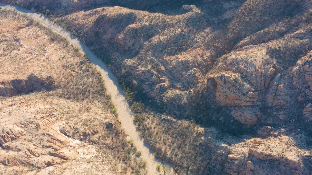 A high aerial view in Cabo Pulmo shows the road curving along the left side of rocky hills, with strong shadow across the canyon and no visible people.