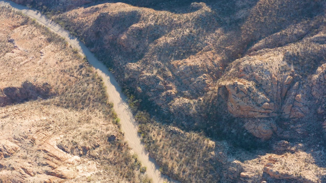 At 0:03, the aerial angle shows the road bending through tan Cabo Pulmo hills while darker rock faces and scrub fill the right side, with no visible people.