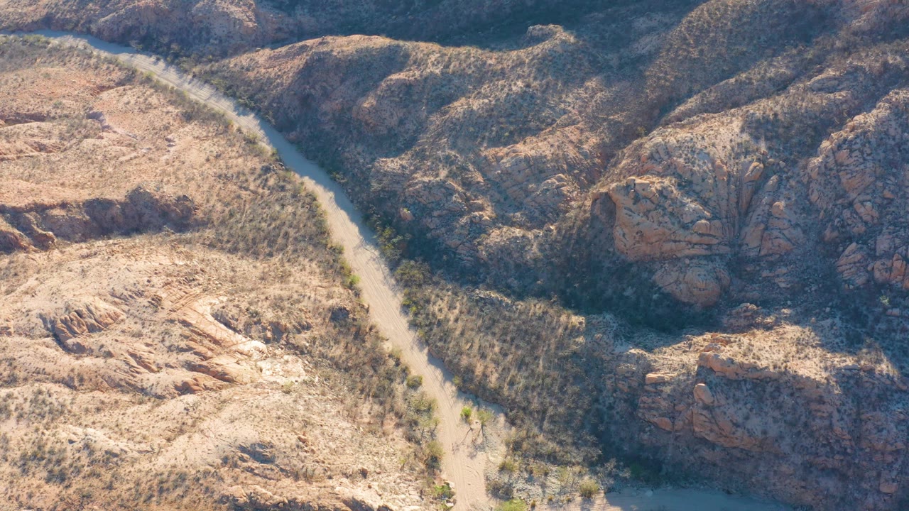 Aerial frame over Cabo Pulmo with the river running along the left edge, a scrub-filled corridor in the center, and layered brown rock ridges on the right, no visible people.