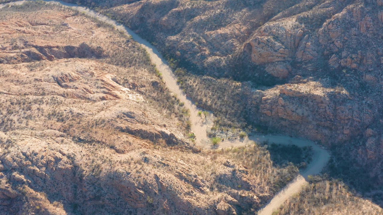Aerial frame over Cabo Pulmo, Baja California Sur showing the winding road beside a darker rocky ridge and sunlit scrub-covered ground with no visible people.