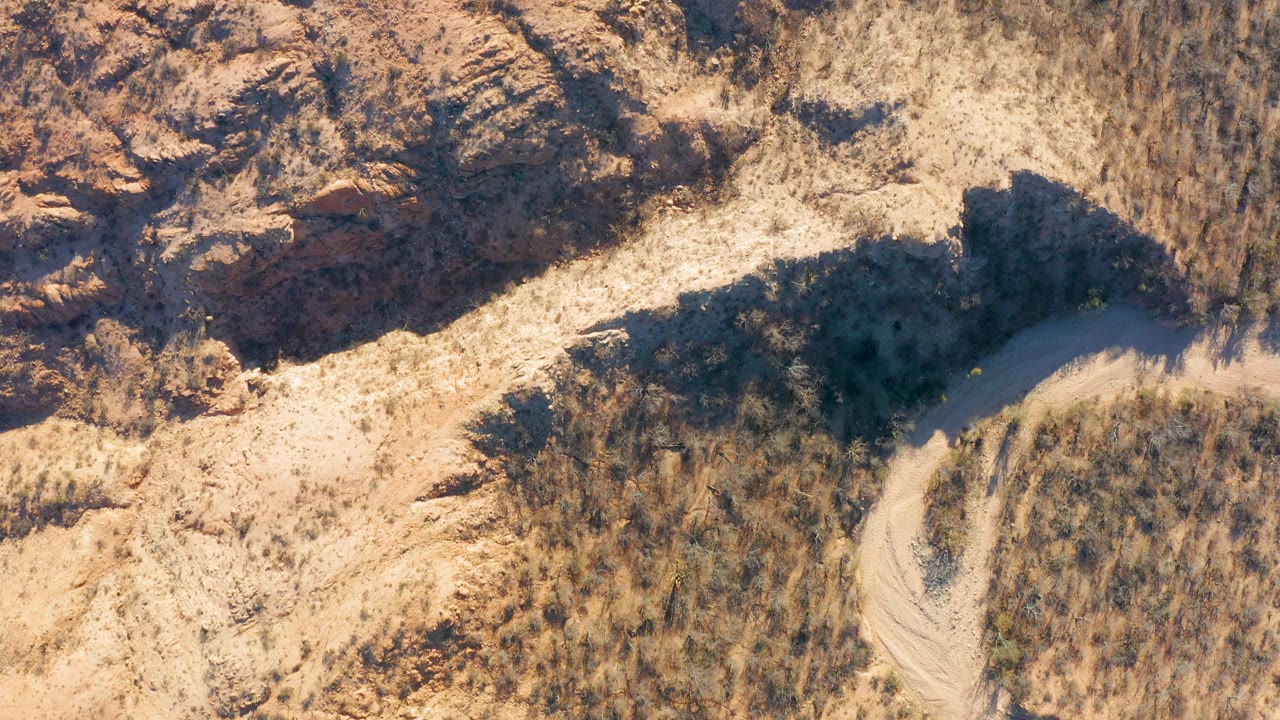 Overhead frame from Cabo Pulmo, Baja California Sur showing a pale winding dirt path beside dry grass and reddish rock formations, with no visible people.