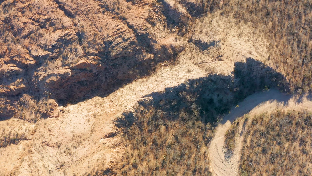 High overhead view in Cabo Pulmo, Baja California Sur with tan desert ground, a partly visible dirt path, dry grasses, and a rocky outcrop casting a dark shadow, with no visible people.
